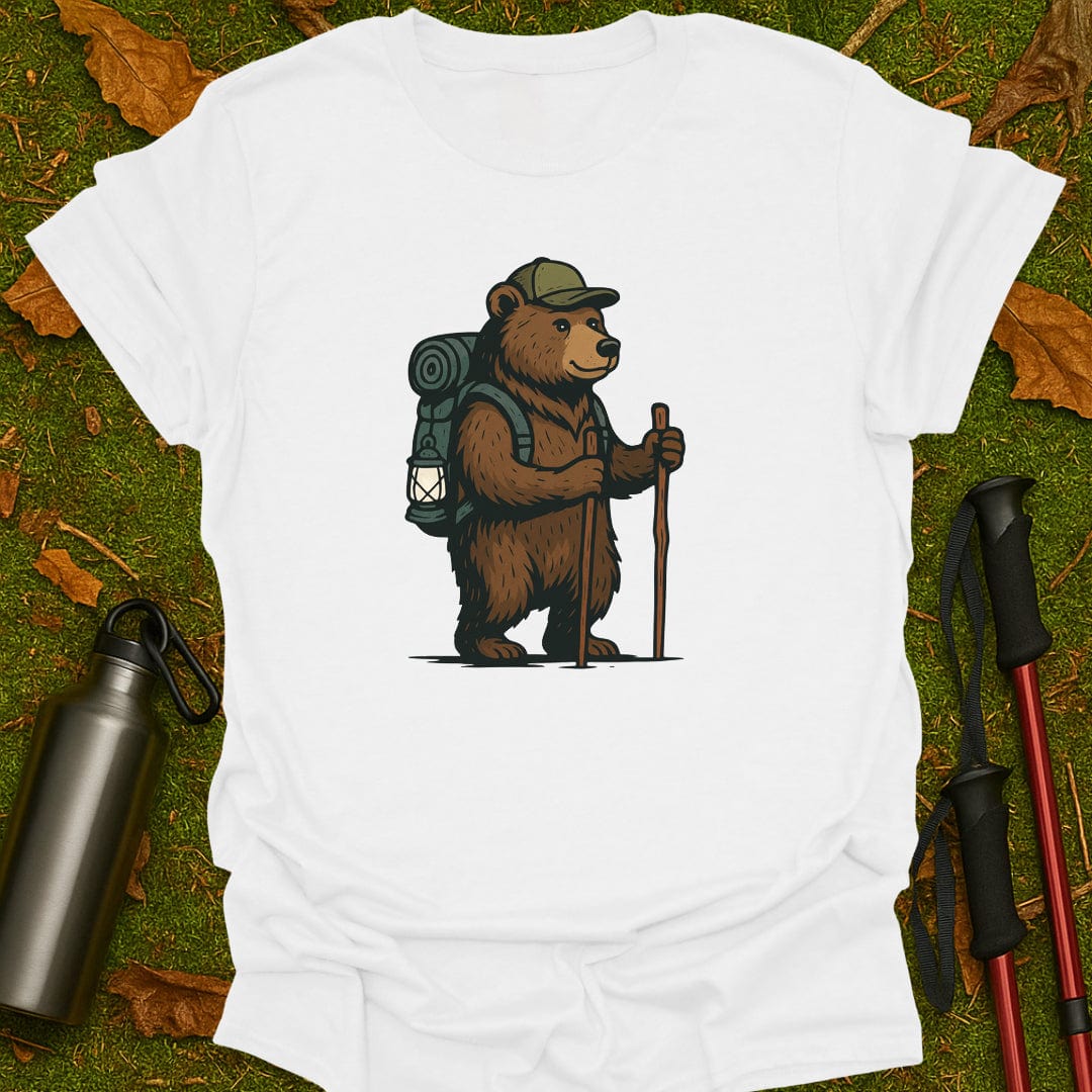 White t-shirt with a bear graphic holding a lantern and walking stick, placed on grass with hiking equipment.